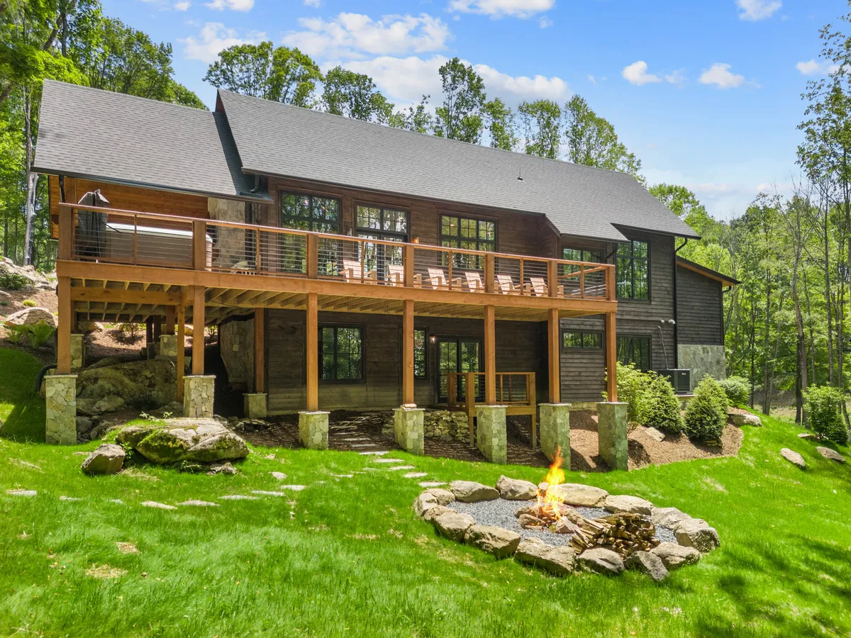 The image showcases the exterior of a luxury mountain cabin in Banner Elk, North Carolina. It highlights a spacious wooden deck with comfortable seating around a fire pit, overlooking a lush green lawn. Large windows reveal the inviting interior, and the scene suggests the warmth of gathering outdoors, with the promise of sunset views and cozy evenings by the fire.