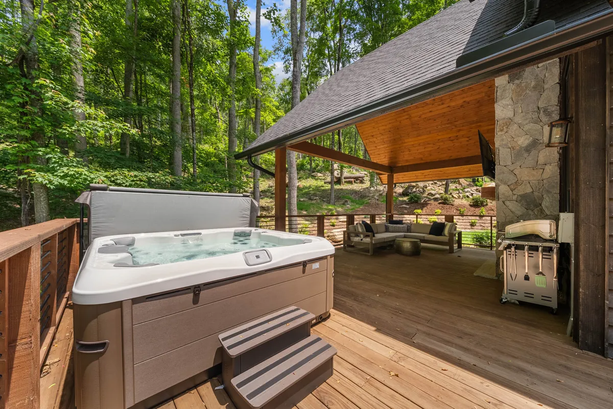 A spacious wooden deck features a hot tub and outdoor seating area nestled among the trees. The serene setting is designed for unwinding, socializing, or enjoying nature, with the deck serving as a bridge between indoor comfort and outdoor adventure.