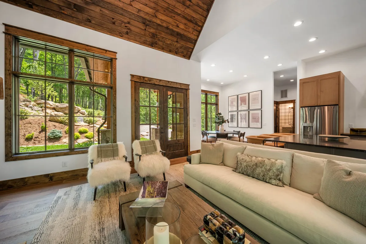This inviting great room features expansive windows framing lush greenery beyond, a plush sofa set with decorative pillows, rustic wooden beams overhead, and a modern kitchen area in the background. The space is designed for both relaxation and socializing, with ample natural light and comfortable furnishings that encourage gathering with family and friends.
