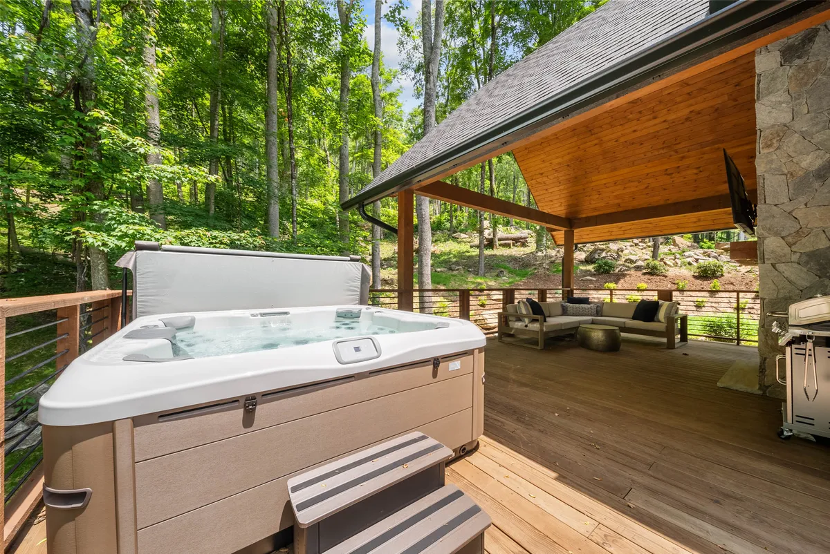 This exterior image captures a large, covered deck area surrounded by forest, featuring a bubbling hot tub, outdoor seating, and ample space for relaxing. The view hints at sunset or evening, inviting guests to soak under the stars or gather with friends.
