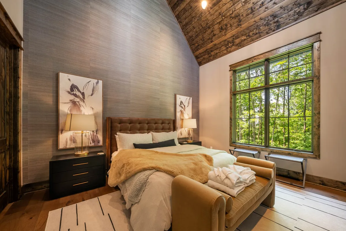A serene bedroom with a comfortable bed draped in a plush throw blanket is bathed in natural light from a large window. Art pieces adorn the walls, and the atmosphere suggests a cozy, restful retreat perfect for unwinding after a day of adventure.