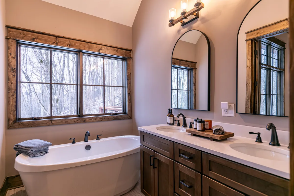 A luxurious bathroom featuring a freestanding soaking tub, a double sink vanity, large windows with woodland views, and warm wooden accents. The space feels spa-like and inviting, ideal for relaxation and self-care.