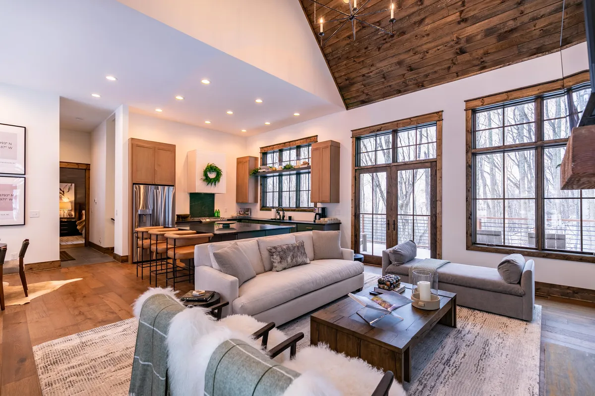 A seamless open-plan space with a comfortable sofa, kitchen island, large windows, and an adjacent dining area. Warm natural light fills the room, inviting guests to relax or socialize. The soft furnishings and layout promote togetherness.