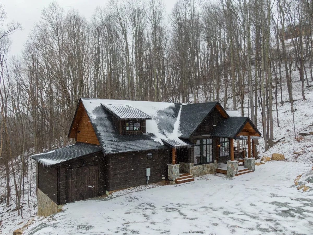 A picturesque exterior view with a snow-covered roof, distinctive wooden and stone details, large windows, and a deck with steps leading to an open area. The surroundings evoke a welcoming, adventure-ready vibe for group escapes.