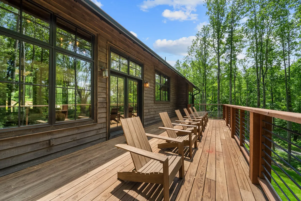 This image presents a spacious wooden deck with Adirondack chairs, large windows, and a backdrop of surrounding trees. The inviting setup encourages relaxation and socializing while connecting seamlessly to the natural woodland setting.