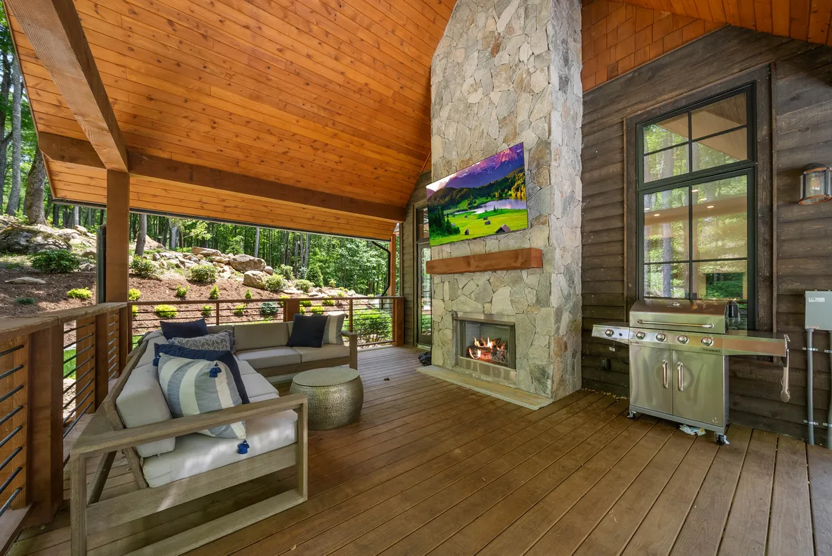 A welcoming outdoor living space is pictured, centered around a large stone gas fireplace, a grill, and plenty of comfortable seating. An outdoor TV is visible above the fireplace, and the seating area opens to nature, with sunset light flooding the scene.