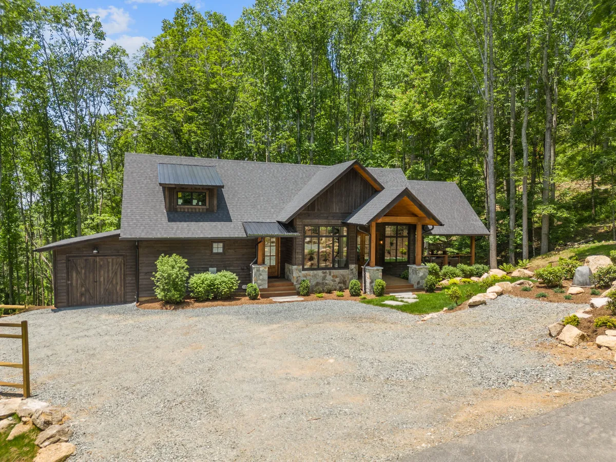 The image features the home's wooden and stone facade, large windows, and a welcoming front porch with steps. It is framed by natural greenery, suggesting a seamless harmony between the house and its mountain environment, and alluding to the exploration that awaits just beyond the doorstep.